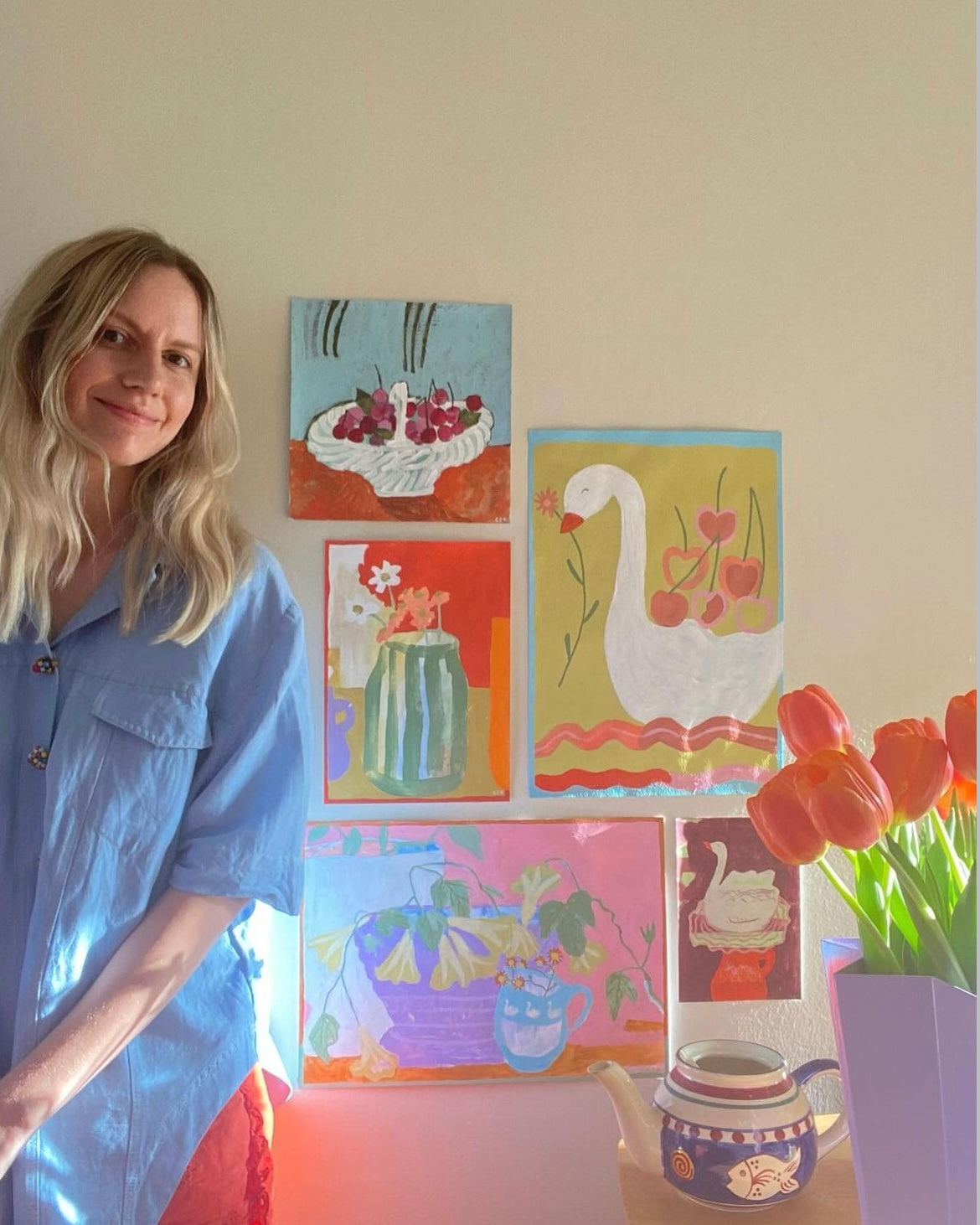 Crystal sitting in her studio next to 5 paintings of various subject matters