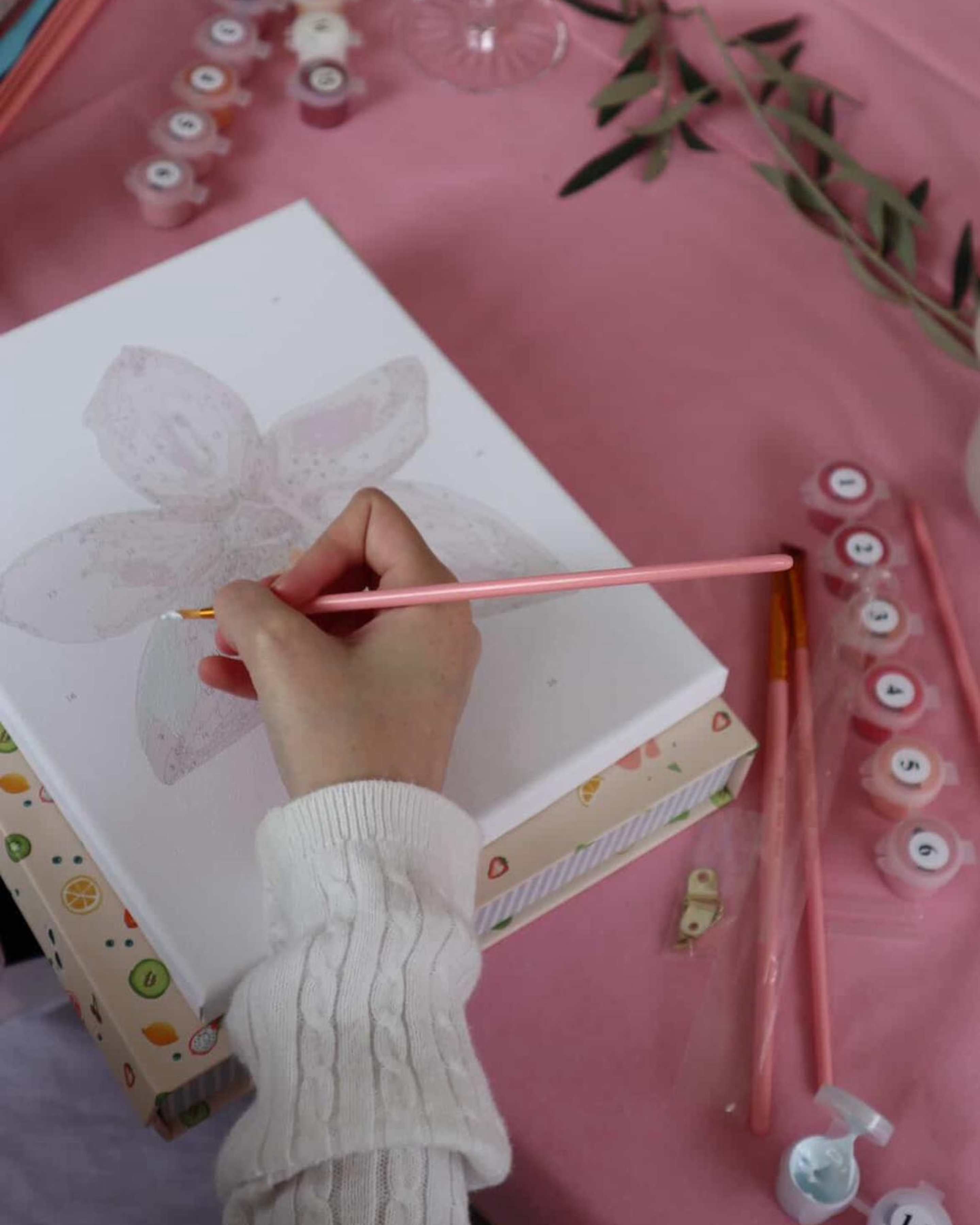 Person painting an orchid flower on a mini framed paint-by-numbers canvas from Paint Like Frida, with pink brush and pastel paints on a pink tablecloth.
