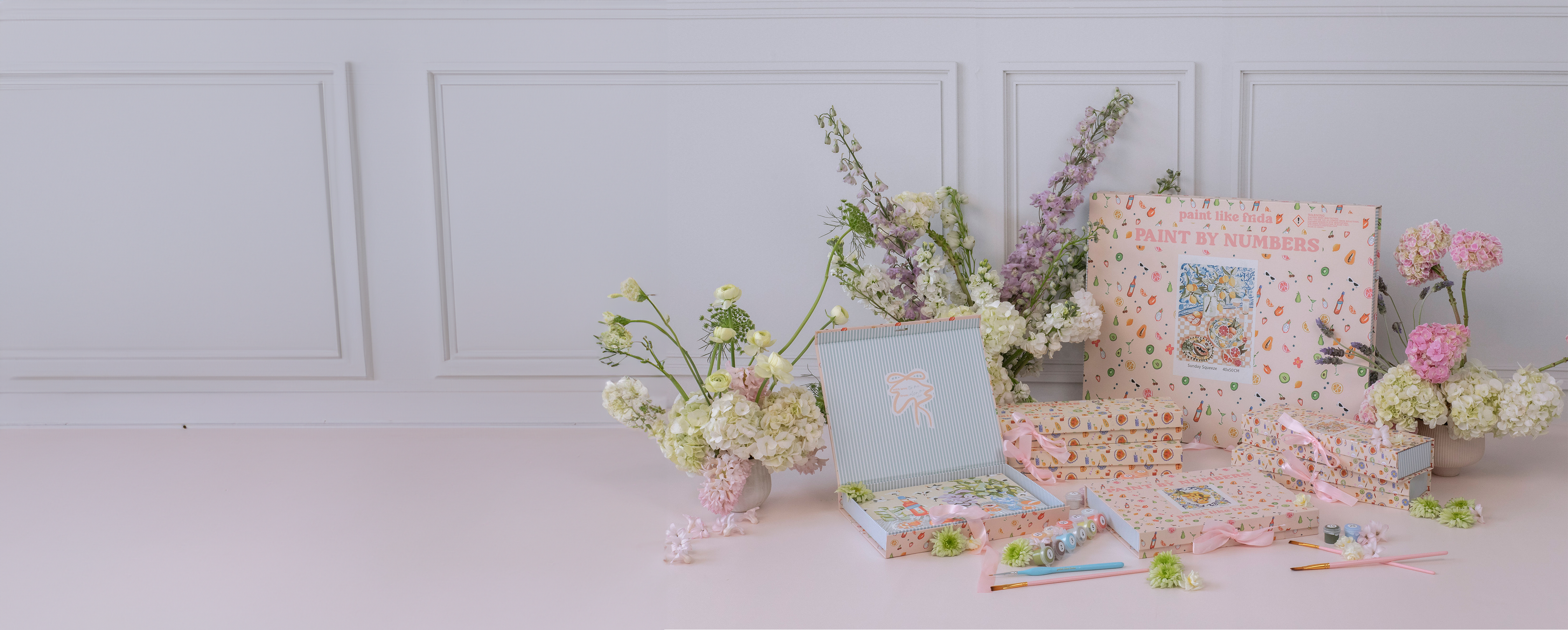 A beautifully styled display of 'Paint Like Frida' paint-by-numbers kits, arranged with pastel-colored packaging featuring illustrated fruit and art supplies. The set includes multiple kits, some tied with soft pink ribbons, along with small paint pots and brushes. Fresh floral arrangements of hydrangeas, stock, and ranunculus in soft pink, white, and purple hues surround the kits, complementing the elegant and feminine aesthetic. The background features a white paneled wall