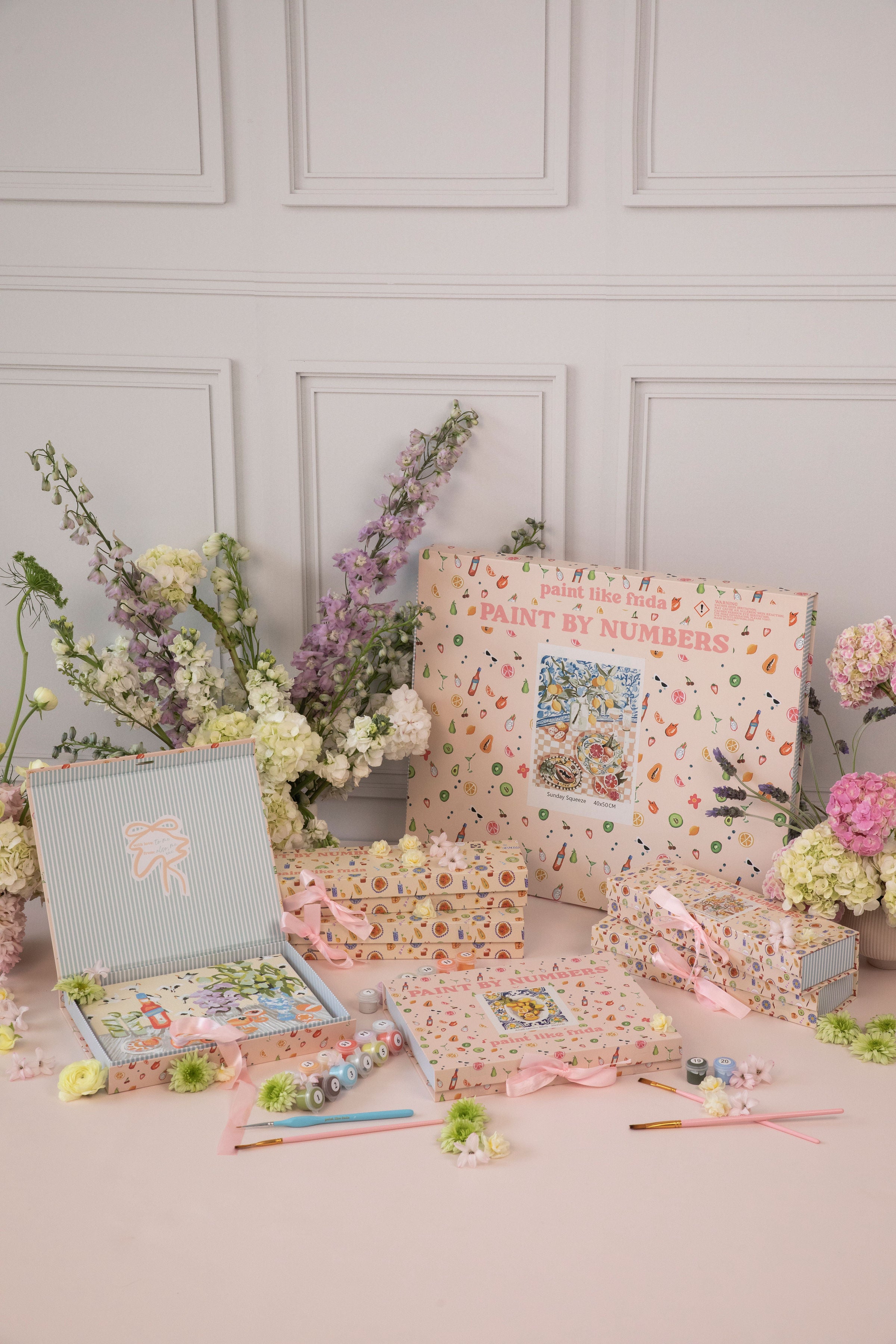 A beautifully styled display of 'Paint Like Frida' paint-by-numbers kits, arranged with pastel-colored packaging featuring illustrated fruit and art supplies. The set includes multiple kits, some tied with soft pink ribbons, along with small paint pots and brushes. Fresh floral arrangements of hydrangeas, stock, and ranunculus in soft pink, white, and purple hues surround the kits, complementing the elegant and feminine aesthetic. The background features a white paneled wall
